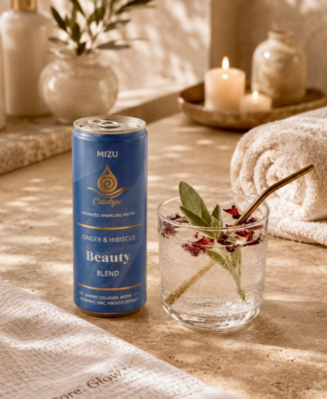 A can of Mizu Cosmetics Beauty Blend serum stands on a stone surface beside a glass of water with green leaves and small red flowers, with folded towels, candles, and bathroom accessories in a warm, sunlit setting.